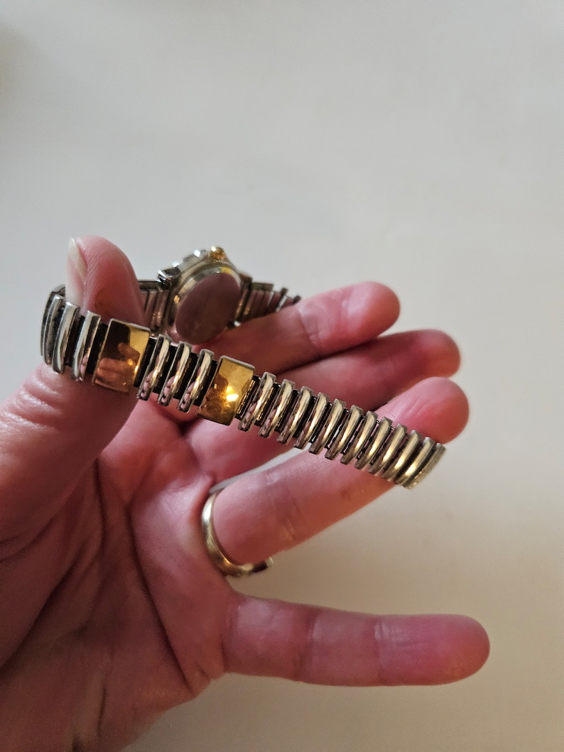 May include: A silver and gold tone watch with a metal band. The watch has a round face and a gold tone bezel.