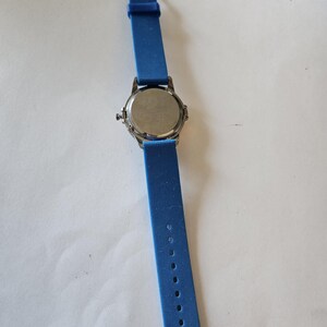 Puede incluir: Un reloj de pulsera con una correa de silicona azul y una esfera plateada. La correa del reloj est&aacute; extendida, mostrando los agujeros de ajuste. El reloj est&aacute; sobre una superficie blanca.