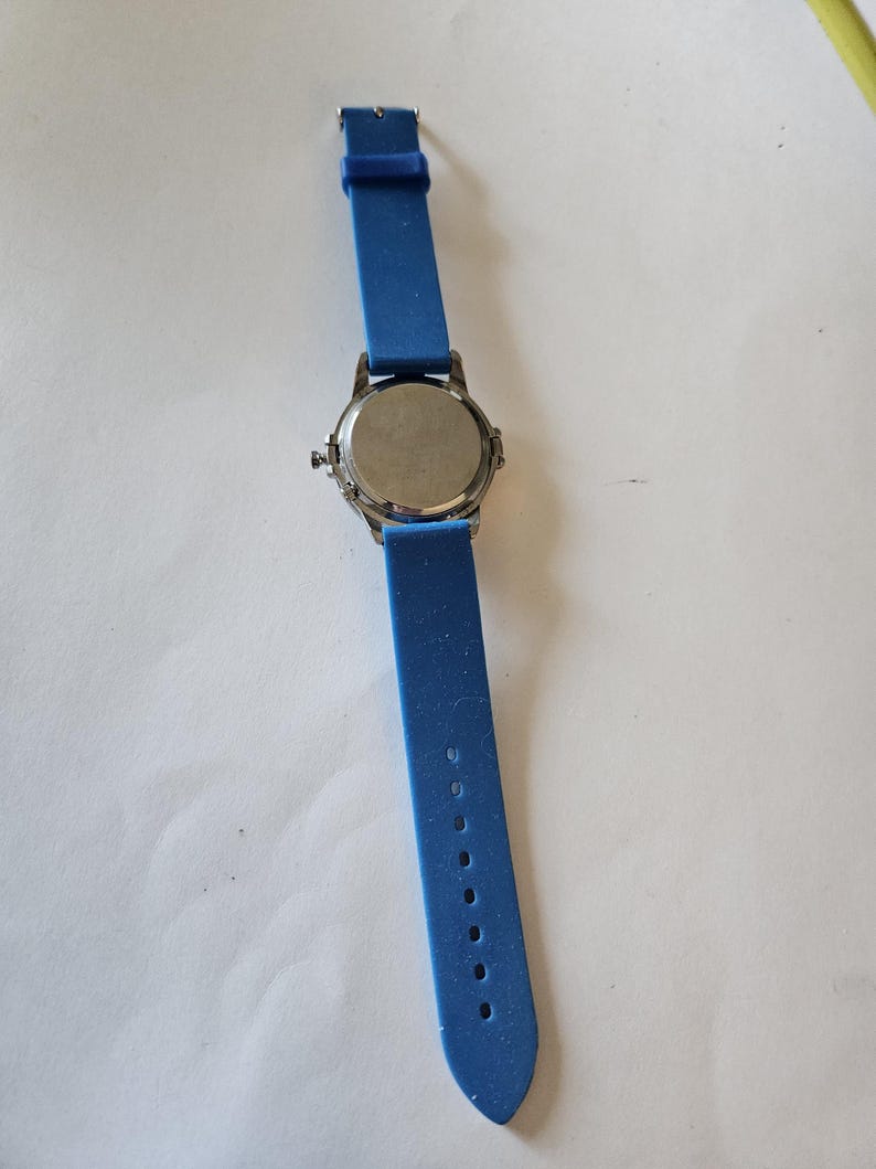 Puede incluir: Un reloj de pulsera con una correa de silicona azul y una esfera plateada. La correa del reloj est&aacute; extendida, mostrando los agujeros de ajuste. El reloj est&aacute; sobre una superficie blanca.