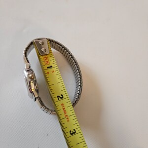 May include: A silver watch with a metal band being measured with a yellow tape measure. The tape measure reads 2 inches.