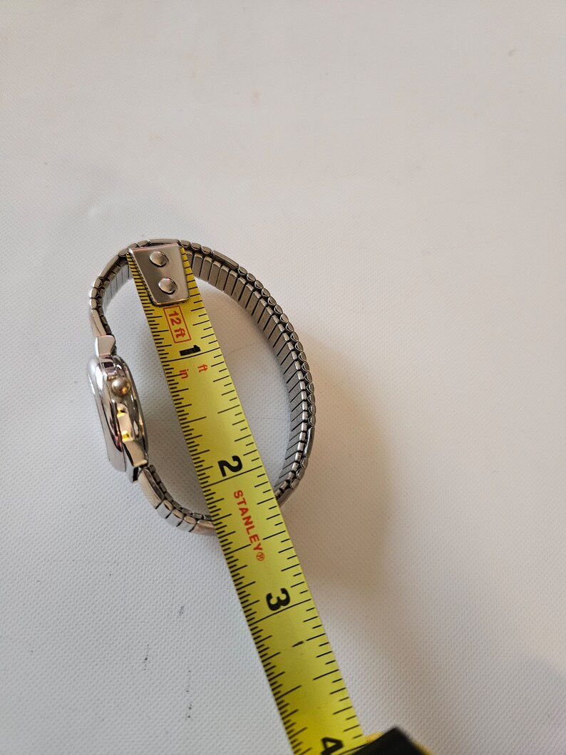 May include: A silver watch with a metal band being measured with a yellow tape measure. The tape measure reads 2 inches.
