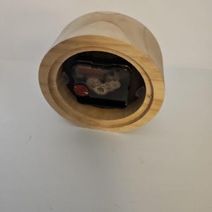 Puede incluir: Un reloj de madera de color marr&oacute;n claro. El reloj tiene forma cil&iacute;ndrica y un mecanismo de reloj visible. La esfera del reloj no es visible. El reloj est&aacute; hecho de madera y tiene un veteado natural.