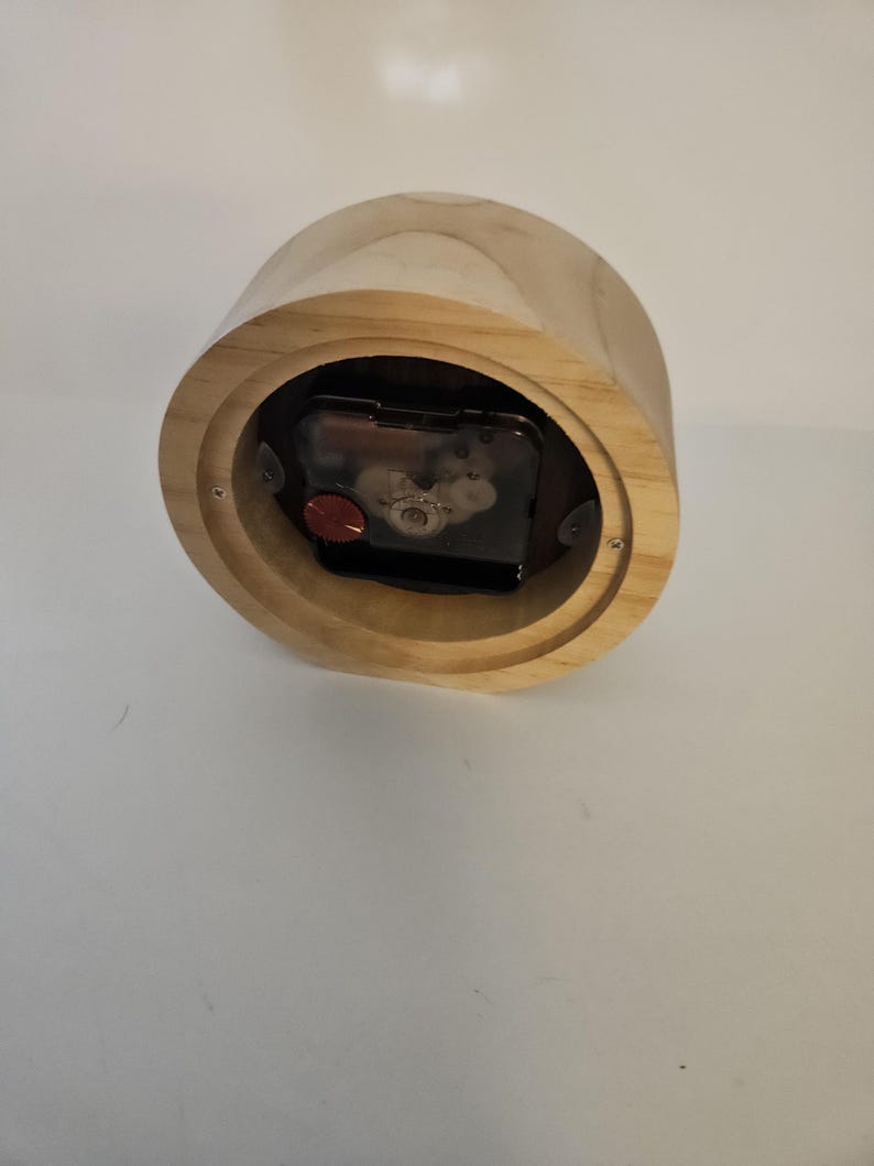 Puede incluir: Un reloj de madera de color marr&oacute;n claro. El reloj tiene forma cil&iacute;ndrica y un mecanismo de reloj visible. La esfera del reloj no es visible. El reloj est&aacute; hecho de madera y tiene un veteado natural.