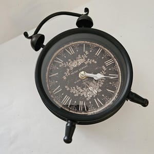 May include: A black metal alarm clock with a round face featuring white Roman numerals and a dark background. The clock face displays the text Henri Dupont and 60 Rue Paris, along with decorative floral illustrations and two white clock hands.