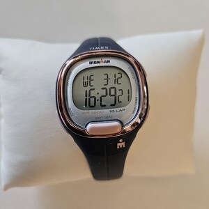 May include: A black and rose gold Ironman watch with a digital display showing the time, date, and lap count. The watch is water resistant to 100 meters.
