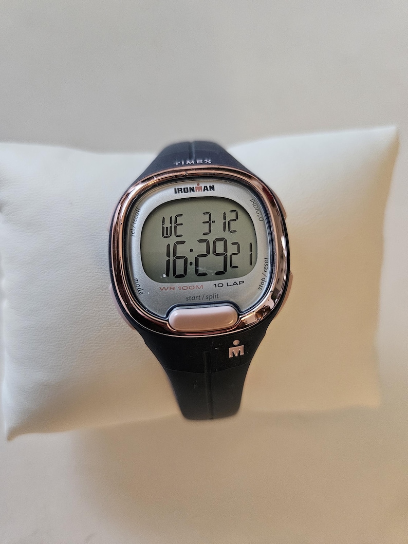 May include: A black and rose gold Ironman watch with a digital display showing the time, date, and lap count. The watch is water resistant to 100 meters.