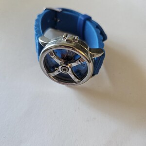 Puede incluir: Un reloj de pulsera azul con esfera plateada y correa de silicona azul. La esfera del reloj tiene un dise&ntilde;o &uacute;nico con un elemento central y un patr&oacute;n en forma de cruz. La correa se asegura con una hebilla plateada.