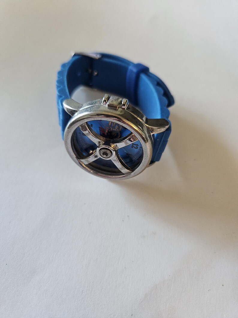 Puede incluir: Un reloj de pulsera azul con esfera plateada y correa de silicona azul. La esfera del reloj tiene un dise&ntilde;o &uacute;nico con un elemento central y un patr&oacute;n en forma de cruz. La correa se asegura con una hebilla plateada.
