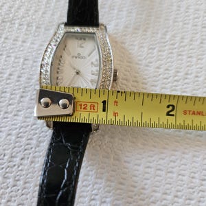 May include: A rectangular wristwatch with a silver face and a black leather strap. The watch face has the brand name "minico" and is surrounded by small, clear crystals. The watch is approximately 2.5 cm wide.