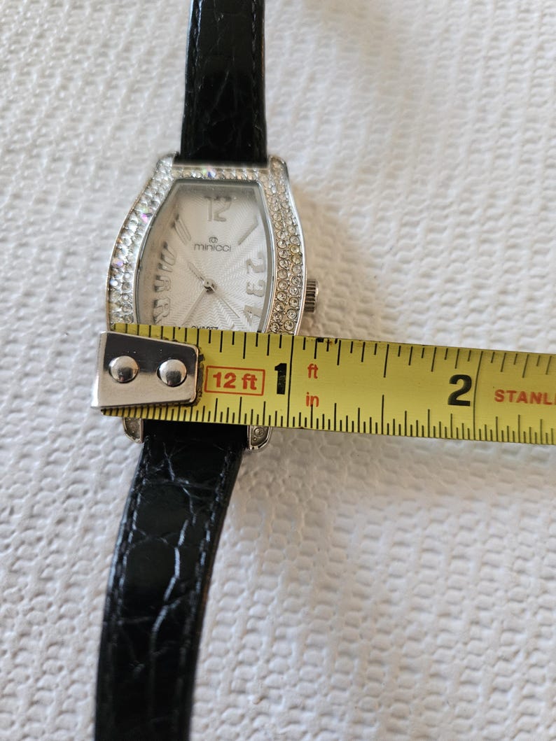 May include: A rectangular wristwatch with a silver face and a black leather strap. The watch face has the brand name "minico" and is surrounded by small, clear crystals. The watch is approximately 2.5 cm wide.