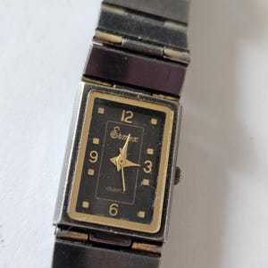 May include: A rectangular Ermex quartz watch with a black face, gold-colored trim, and silver-tone metal band. The watch face displays the numbers 3, 6, 9, and 12, with gold-colored hands. The watch band is made of multiple rectangular links.