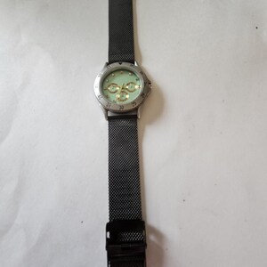 May include: A wristwatch featuring a black mesh metal strap and a silver-tone circular case. The watch face is a pale mint green color with three gold-tone sub-dials and gold-tone hour markers. The bezel is silver with black numerical markings.