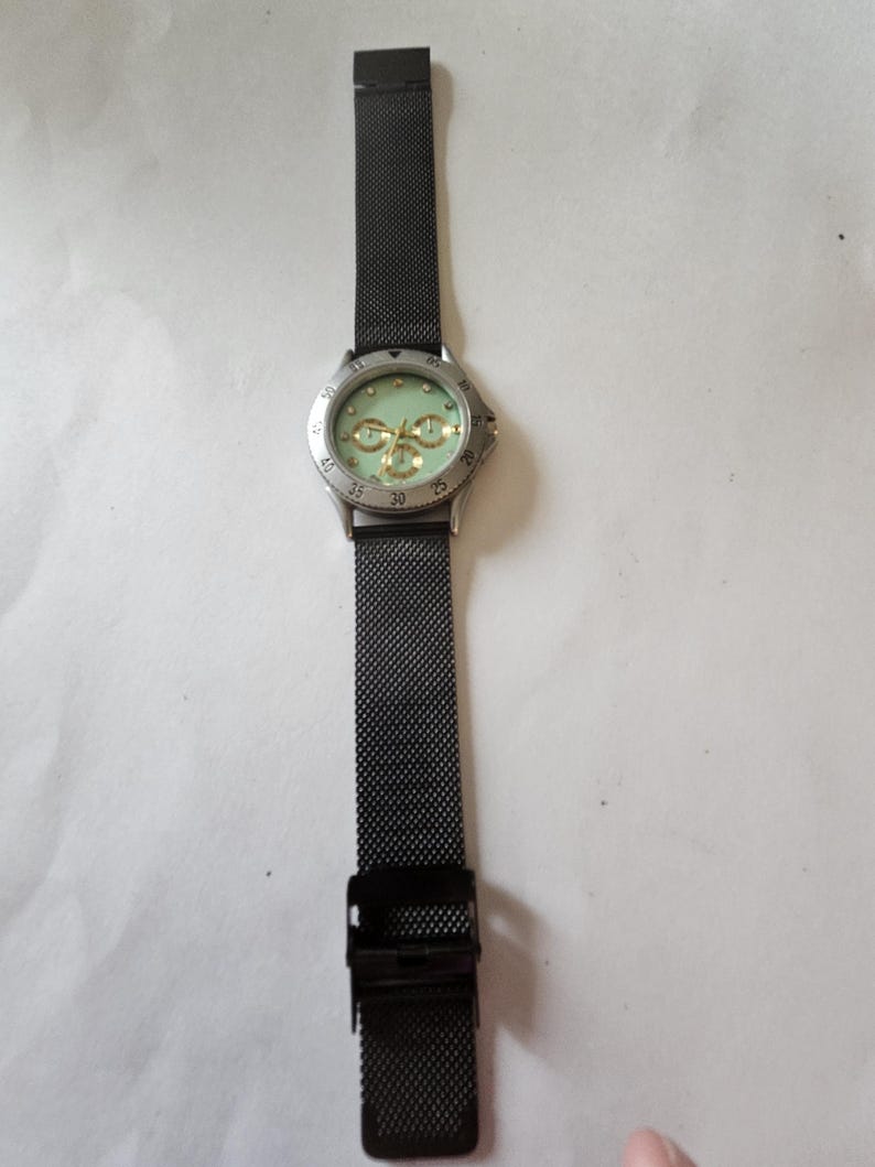 May include: A wristwatch featuring a black mesh metal strap and a silver-tone circular case. The watch face is a pale mint green color with three gold-tone sub-dials and gold-tone hour markers. The bezel is silver with black numerical markings.