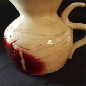 May include: A white ceramic pitcher with a red and white glaze. The pitcher has a unique handle and a textured surface.