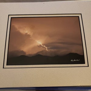 May include: A framed photograph of a lightning strike over a mountain range. The sky is a warm orange color and the lightning is a bright white. The photograph is signed "Tony  B. Wilson".