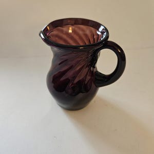 May include: A small, dark purple glass pitcher with a handle. The pitcher has a flared rim and a swirling design on the body. The handle is a solid, rounded shape. The pitcher is sitting on a white surface.
