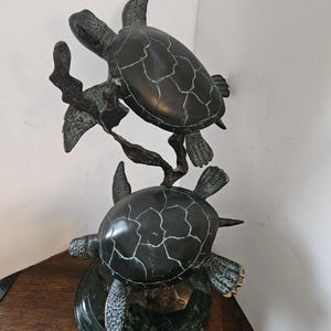 May include: A bronze sculpture of two sea turtles, one atop the other, with a dark green patina. The turtles have detailed shells and flippers, and are mounted on a green marble base. The sculpture is a decorative item.