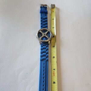 Puede incluir: Un reloj de pulsera azul con esfera plateada y correa de silicona azul. El reloj est&aacute; junto a una cinta m&eacute;trica amarilla, que muestra que la correa mide aproximadamente 23 cm.