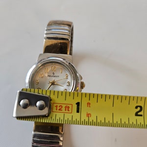 May include: A silver and gold two-tone wristwatch with a white face and black numbers. The watch is 1 inch in diameter and has a Peugeot logo on the face.