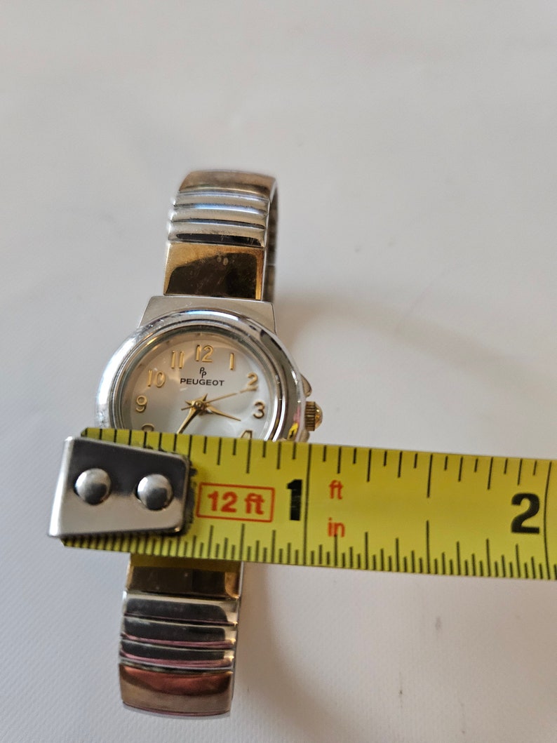 May include: A silver and gold two-tone wristwatch with a white face and black numbers. The watch is 1 inch in diameter and has a Peugeot logo on the face.