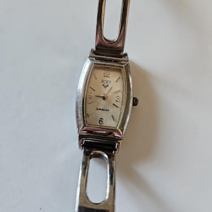May include: A silver Roxy Quiksilver wristwatch with a rectangular face. The watch face is cream-colored with black numerals and hands. The watch band is silver and has an open design.