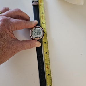 May include: A black digital watch with a rose gold trim and a black rubber band. The watch face displays the time, date, and day of the week. The watch is measured against a yellow tape measure.