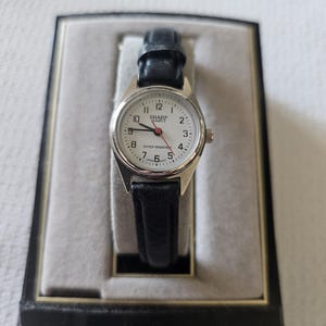 May include: A silver-toned Sharp Quartz wristwatch with a white face, black numerals, and a black leather band. The watch is displayed in a black box with a gray interior. The watch face reads "SHARP QUARTZ" and "WATER RESISTANT."