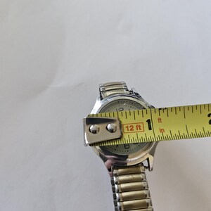 May include: Silver-toned wristwatch with a light-colored face, featuring black numerals and hands. The watch is being measured with a yellow tape measure, showing a width of approximately 1 inch. The watch band is made of silver-colored metal links.