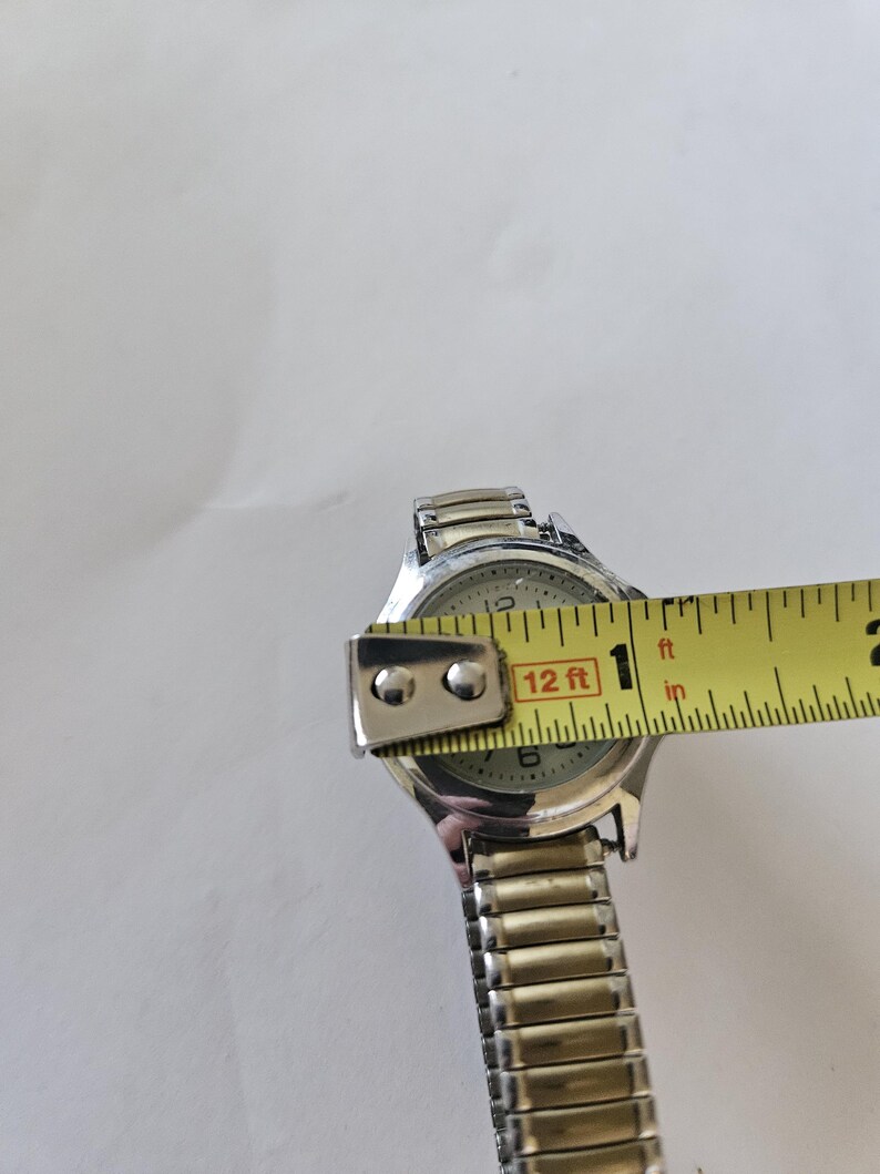 May include: Silver-toned wristwatch with a light-colored face, featuring black numerals and hands. The watch is being measured with a yellow tape measure, showing a width of approximately 1 inch. The watch band is made of silver-colored metal links.