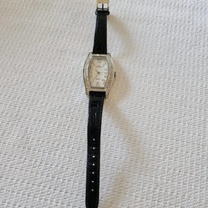 May include: A rectangular silver wristwatch with a white face and a black leather strap. The watch face has silver hands and is surrounded by small, clear crystals. The strap has a buckle closure.