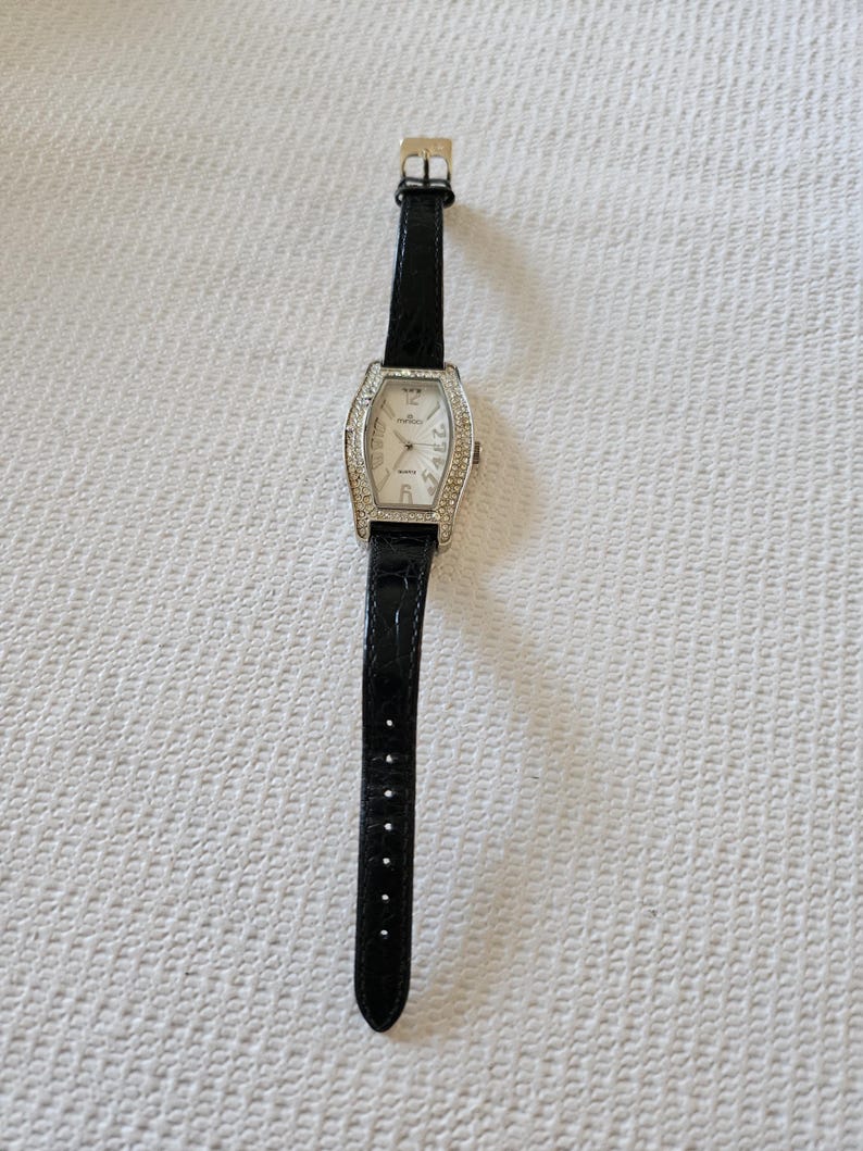 May include: A rectangular silver wristwatch with a white face and a black leather strap. The watch face has silver hands and is surrounded by small, clear crystals. The strap has a buckle closure.