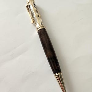 May include: A ballpoint pen featuring a dark brown, polished wooden barrel and polished gold-tone metal components. The pen includes a gold-tone clip, a decorative center band, and a tapered gold-tone tip, showcasing a sleek and functional design.