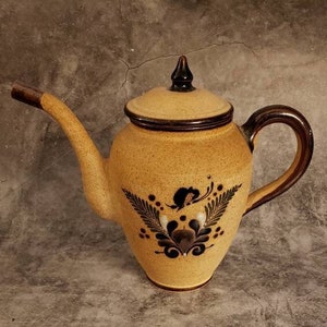 May include: A brown and tan ceramic teapot with a black handle and a floral design. The teapot has a spout and a lid.