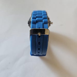 Puede incluir: Un reloj de pulsera azul con un cierre de metal plateado. La correa del reloj es de un material azul texturizado. La esfera del reloj es visible en el fondo.