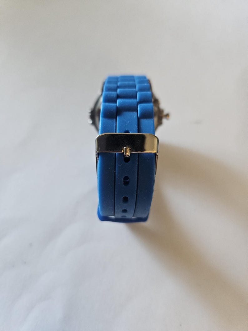 Puede incluir: Un reloj de pulsera azul con un cierre de metal plateado. La correa del reloj es de un material azul texturizado. La esfera del reloj es visible en el fondo.
