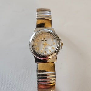 May include: A silver and gold two-tone watch with a white face and black hands. The watch face has gold numbers and the brand name "Peugeot" in gold lettering. The watch has a silver metal band with a gold accent.