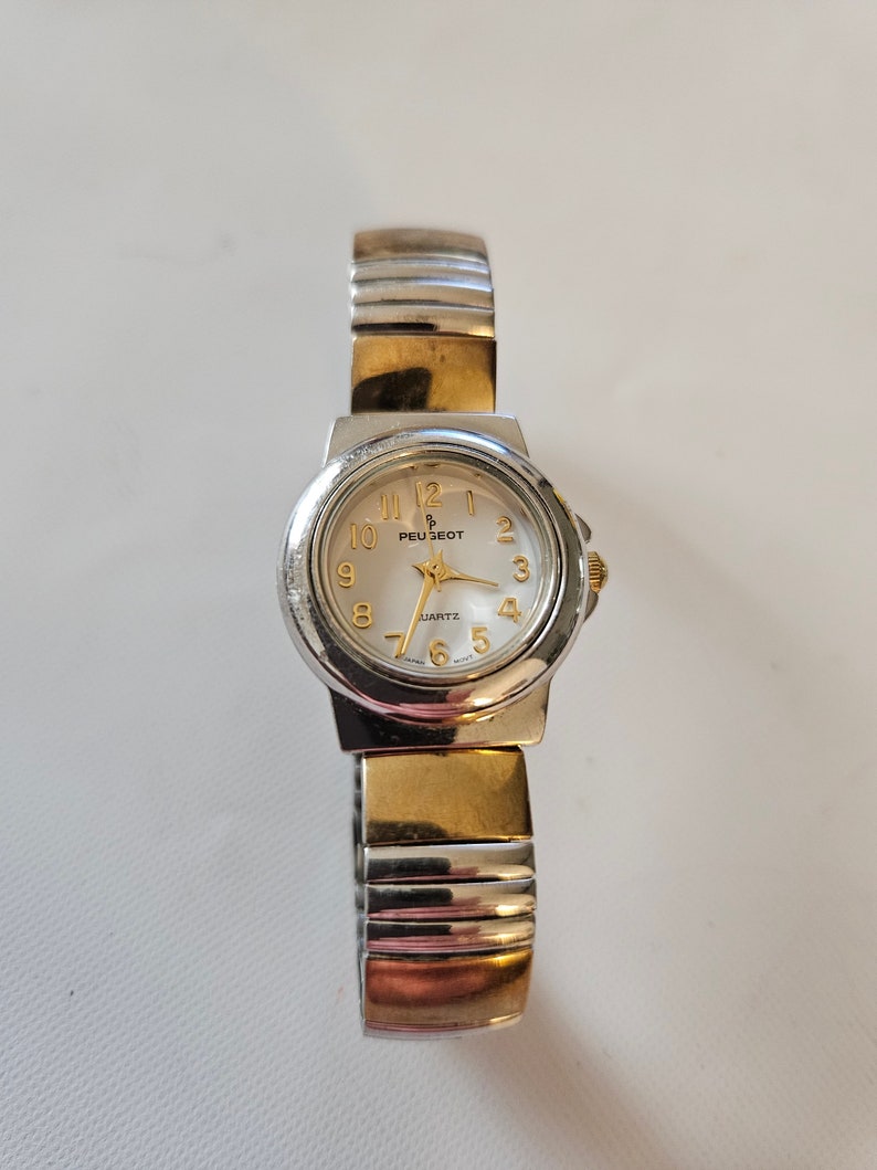 May include: A silver and gold two-tone watch with a white face and black hands. The watch face has gold numbers and the brand name "Peugeot" in gold lettering. The watch has a silver metal band with a gold accent.