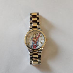 May include: A wristwatch with a gold-tone bezel and a silver-tone band. The watch face features a colorful illustration of a superheroine in a red and blue costume. The watch has gold-tone hands and hour markers.