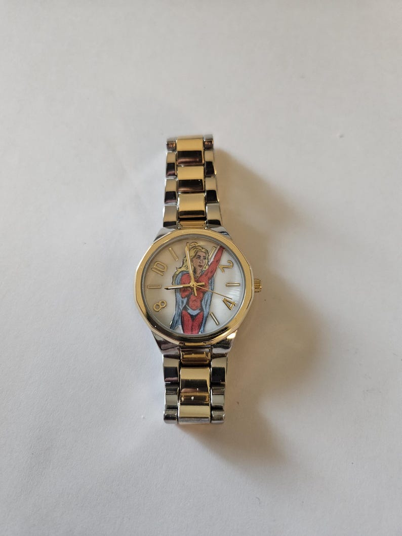 May include: A wristwatch with a gold-tone bezel and a silver-tone band. The watch face features a colorful illustration of a superheroine in a red and blue costume. The watch has gold-tone hands and hour markers.