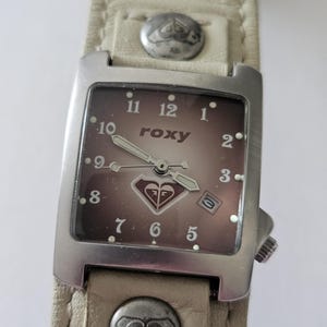 May include: Roxy wristwatch with a square, silver-toned face and a brown dial. The watch features a cream-colored leather band with silver-toned studs. The Roxy logo is visible on the watch face. The watch band measures approximately 1 inch wide.
