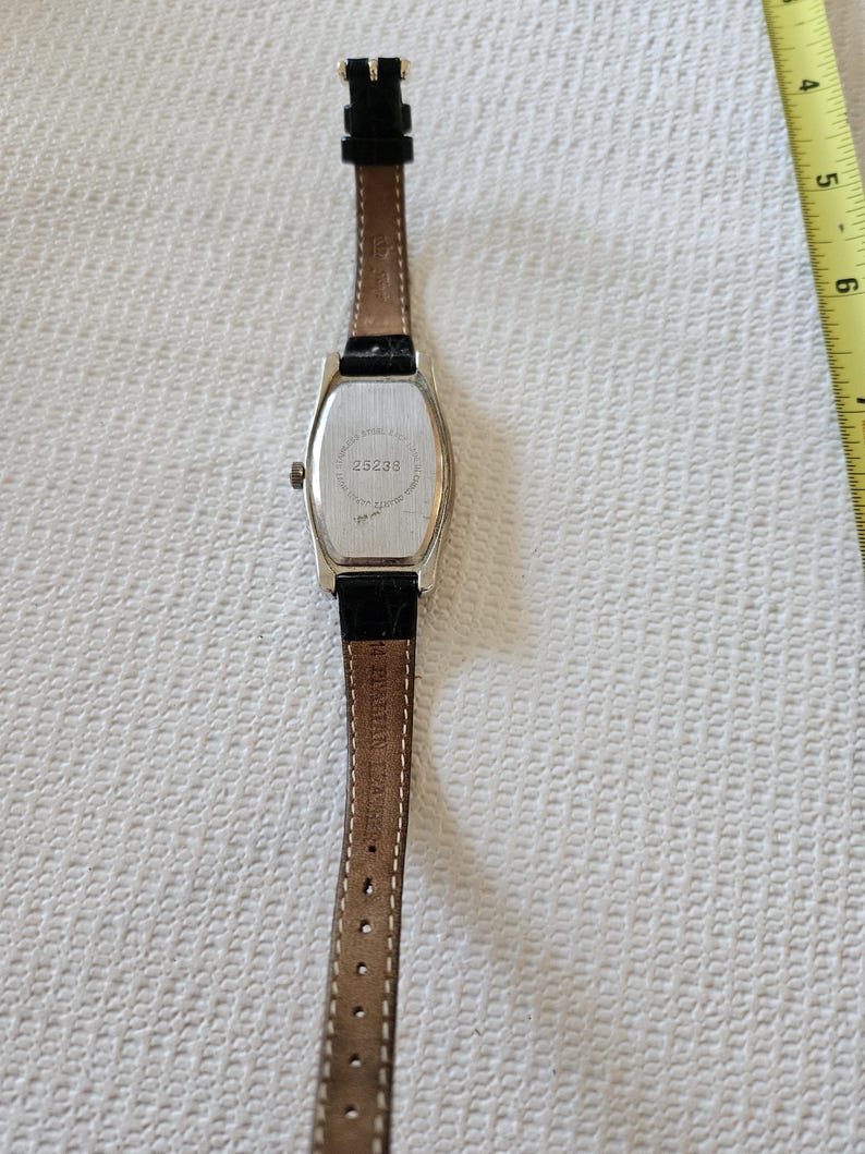 May include: A rectangular silver watch with a black leather strap. The watch face is not visible. The back of the watch has the number "25238" printed on it. The watch strap is brown on the inside.