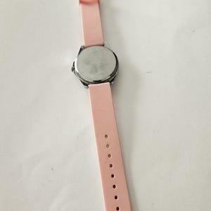 Op de afbeelding: Een polshorloge met een lichtroze siliconen band en een zilverkleurige metalen kast. De horlogeband wordt vastgezet met een zilveren gesp. De wijzerplaat van het horloge is niet zichtbaar op deze afbeelding.