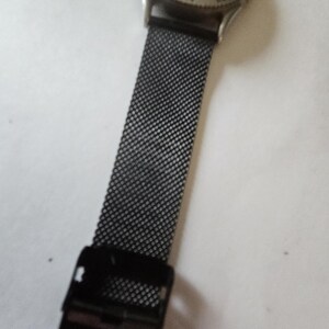 May include: A wristwatch featuring a round silver-tone metal case with a light green dial and a black mesh metal strap. The watch includes a black adjustable sliding clasp mechanism on the band, providing a secure fit for the wrist.