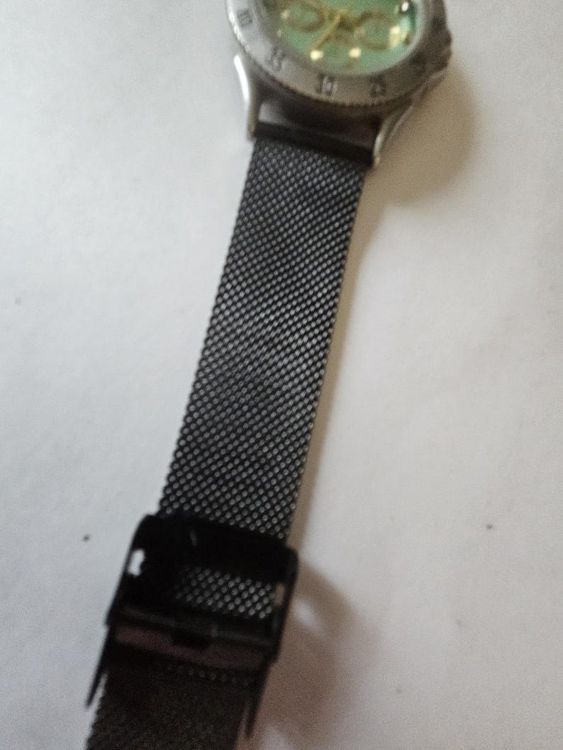 May include: A wristwatch featuring a round silver-tone metal case with a light green dial and a black mesh metal strap. The watch includes a black adjustable sliding clasp mechanism on the band, providing a secure fit for the wrist.