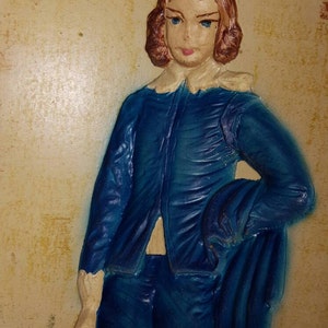 May include: A blue and white figurine of a person with brown hair wearing a blue jacket and pants. The figurine is set against a beige background.
