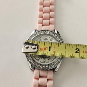 Op de afbeelding: Een lichtroze polshorloge met een zilveren lunette bezet met strass-steentjes. De wijzerplaat is wit met zilveren wijzers en markeringen. De horlogeband is lichtroze. De wijzerplaat heeft een diameter van ongeveer 2,5 cm.