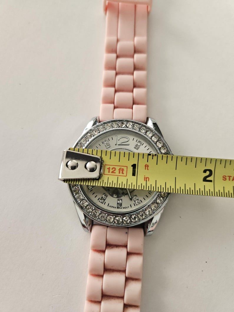 Op de afbeelding: Een lichtroze polshorloge met een zilveren lunette bezet met strass-steentjes. De wijzerplaat is wit met zilveren wijzers en markeringen. De horlogeband is lichtroze. De wijzerplaat heeft een diameter van ongeveer 2,5 cm.