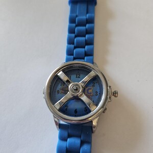 Puede incluir: Un reloj de pulsera azul con bisel plateado y correa azul. La esfera del reloj es azul con la palabra "SONIC" en una fuente estilizada. La correa est&aacute; hecha de un material flexible y texturizado.