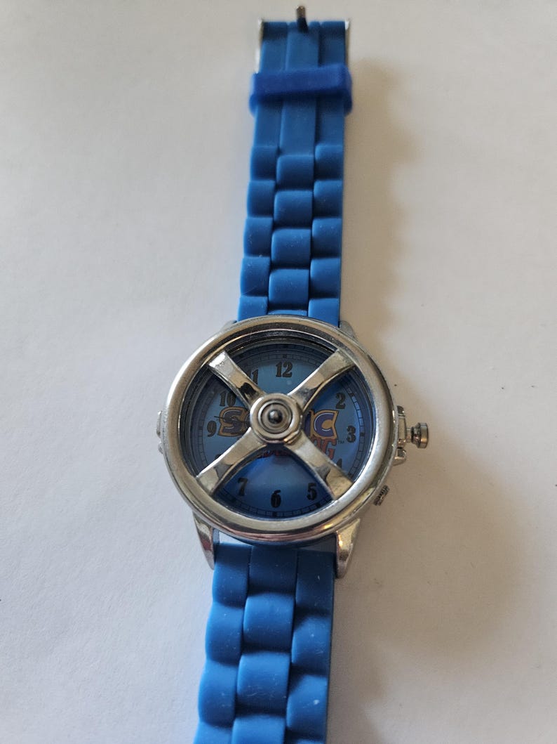 Puede incluir: Un reloj de pulsera azul con bisel plateado y correa azul. La esfera del reloj es azul con la palabra "SONIC" en una fuente estilizada. La correa est&aacute; hecha de un material flexible y texturizado.
