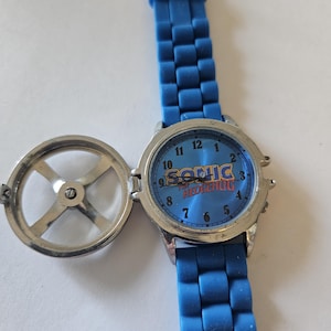 Puede incluir: Un reloj azul de Sonic the Hedgehog con una correa azul a juego. La esfera del reloj presenta el logotipo de Sonic y un fondo azul. El reloj tiene una caja de metal plateado y una tapa con bisagras.